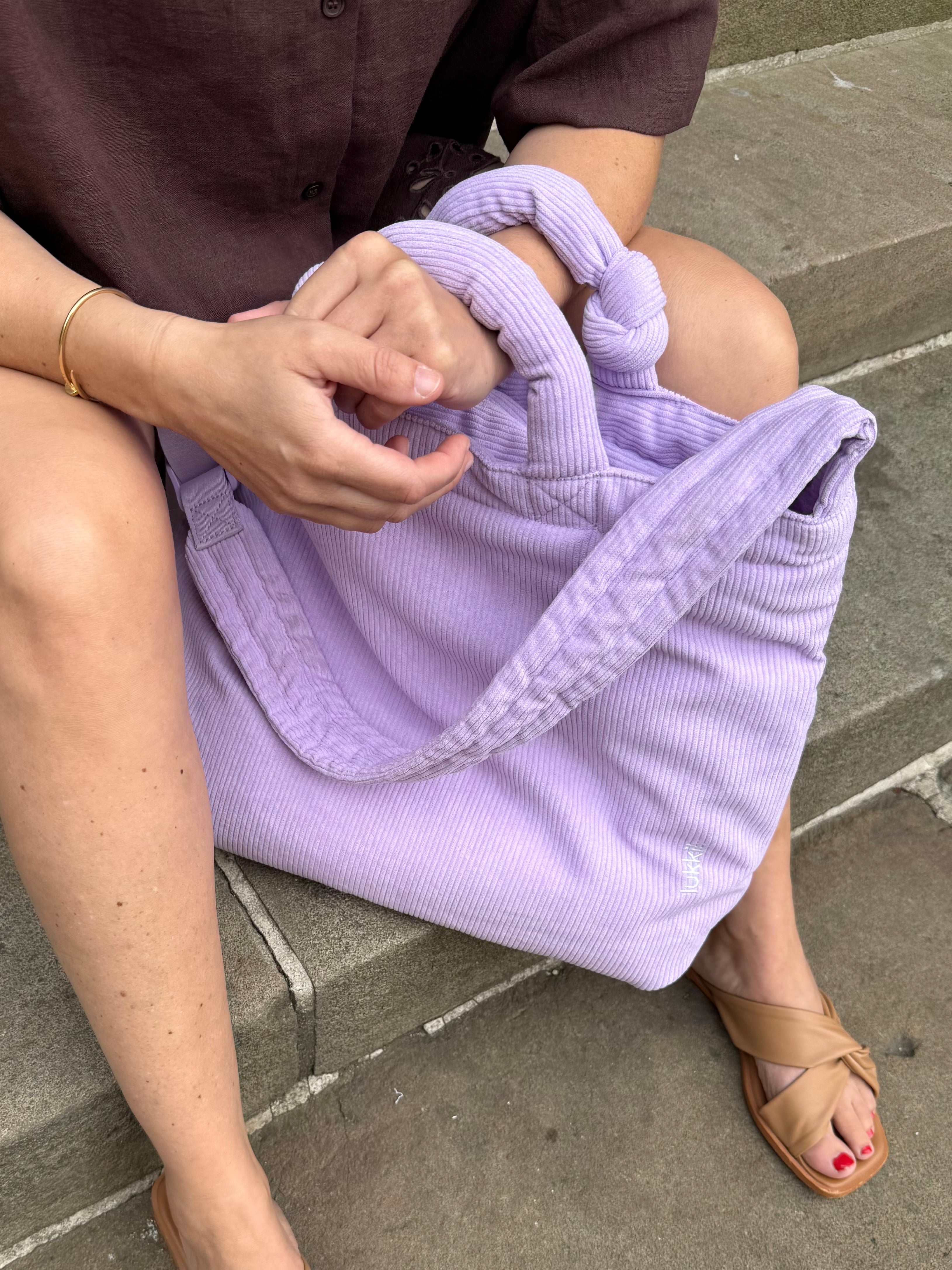 Cord Wickeltasche in Lila mit Knotendetail an den Kurzhenkeln und längenverstellbarem Crossbody-Schultergurt.