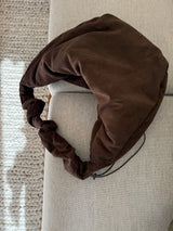 Braune Lukkily Ruffle Bag aus weichem Cord mit breitem Taschenkörper und verspieltem Rüschenhenkel auf hellem Sofa.