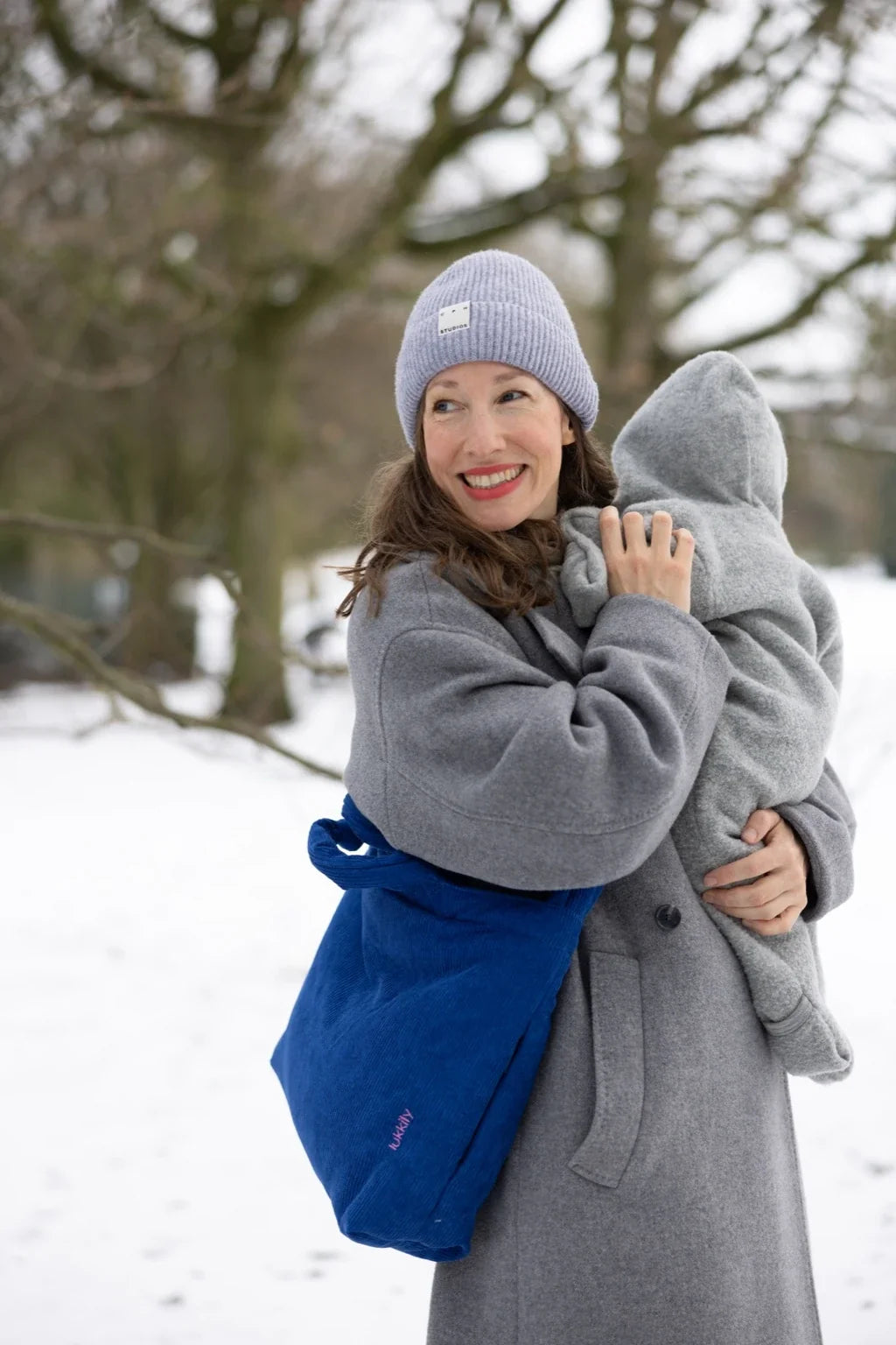 Mama mit blauer Lukkily Wickeltasche in Cordoptik crossbody getragen über einem Mantel mit Baby auf dem Arm