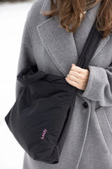 Lukkily Tote Bag Nylon black - hier lässig am gepolsterten, längenverstellbaren Gurt als Crossbodybag über einem Wollmantel getragen, sodass man das pinke Logodetail und das versteckte Außenfach mit Zip sehen kann